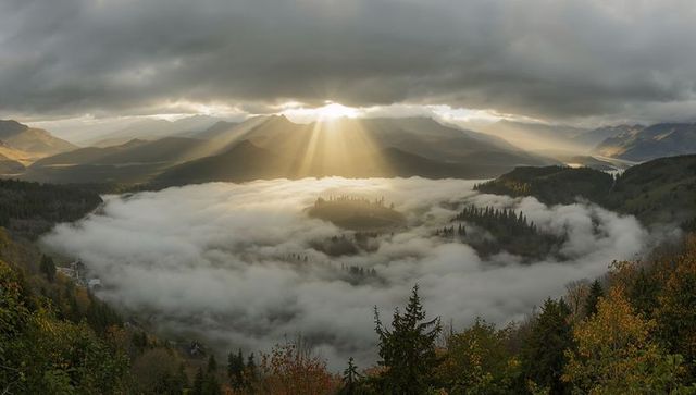 Golden sunbeams piercing cloud gap over fog-filled alpine valley with autumn trees