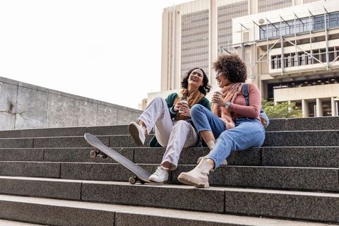 Two friends laughing on campus steps with skateboard and takeaway coffee