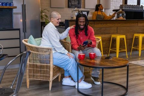 Diverse couple sharing smartphone laughing over coffee in cozy modern cafe lounge