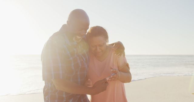 Senior couple laughing and embracing at the beach during sunset. They are looking at something on a mobile phone. This moment captures the joy and bond of elderly relationships, making it perfect for use in advertisements for retirement plans, senior care services, travel agencies, and lifestyle blogs.