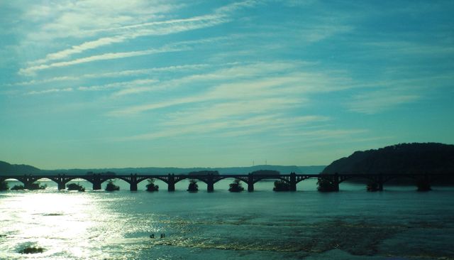 Historic bridge spanning calm river with blue sky and hills in background. Ideal for travel, architecture, landscape, and tourism themes. Perfect for use in promotional materials, travel guides, and nature-focused projects.