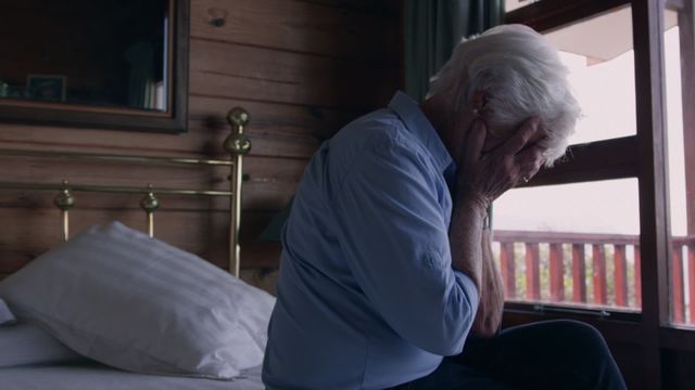 Elderly woman is sitting on a bed, covering her face with her hands, and conveying emotions of sadness and stress possibly related to personal difficulties. Ideal for use in articles or content related to mental health awareness, aging challenges, or senior isolation.