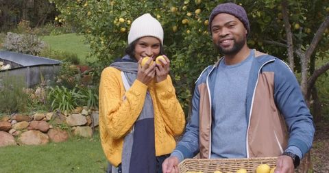 Couple harvesting lemons in backyard orchard with wicker basket wearing cozy knit beanies
