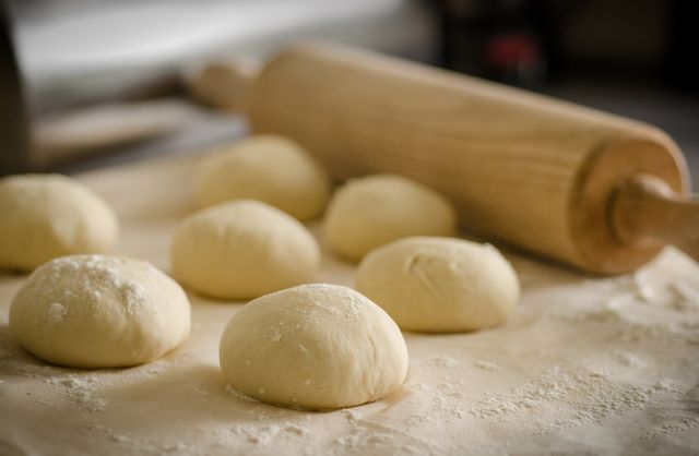 This image shows multiple pizza dough balls and a rolling pin on a wooden surface. Ideal for use in culinary blogs, recipe books, restaurant menus, food advertisements, and kitchen-related content. Perfect for illustrating homemade or restaurant-style pizza preparation.