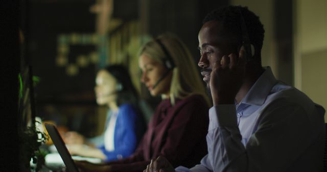 Image of smiling african american businessman and colleagues using headsets at night in office. Business, communication, inclusivity and flexible working concept digitally generated image.