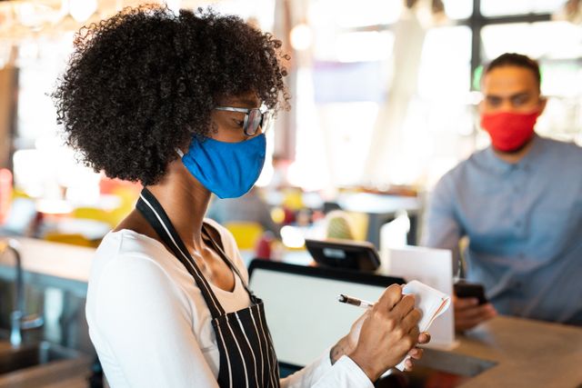 African American female barista wearing face mask taking order from male customer also in face mask. Scene depicts safety measures during COVID-19 pandemic in a coffee shop. Useful for illustrating pandemic-related safety protocols, hospitality industry adaptations, and social distancing practices.