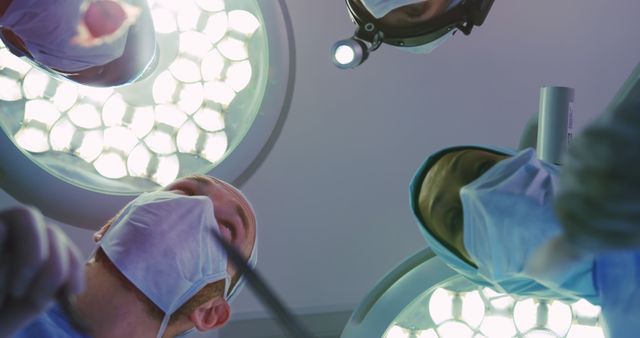 Surgeons actively engaged in a medical procedure, with surgical lights illuminating the scene from above. This visual is perfect for highlighting themes around healthcare, medical education, and hospital environments. Suitable for use in medical journals, healthcare advertising, and educational purposes.