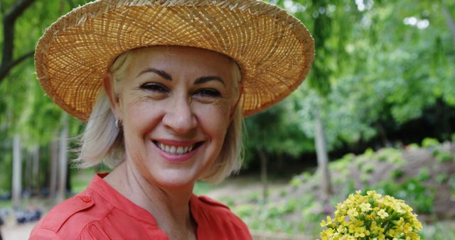 Senior woman wearing a straw hat is smiling while holding a bunch of yellow flowers in a garden. Vibrant greenery and flowering plants are in the background. Suitable for themes related to gardening hobbies, outdoor leisure, senior lifestyle, and enjoying nature.