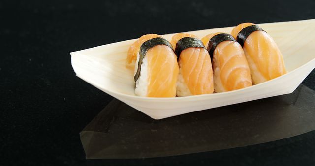A wooden boat-shaped plate holds a row of salmon nigiri sushi, elegantly presented on a dark reflective surface. Salmon nigiri sushi is a popular Japanese dish consisting of a slice of raw salmon atop a mound of vinegared rice, often bound with a strip of seaweed.