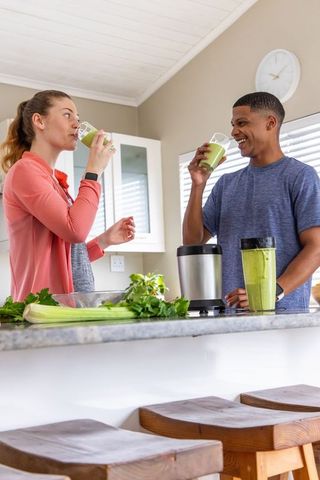 Diverse couple making green smoothies at home kitchen counter with blender, celery