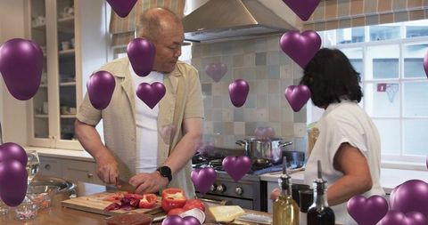 Mature couple cooking together in bright kitchen chopping peppers with floating purple hearts