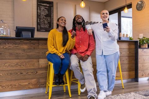 Laughing diverse friends chatting with smartphone at modern cafe counter on yellow stools