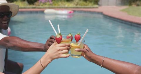 Friends clinking summer cocktails by backyard pool with strawberry and lemon garnish