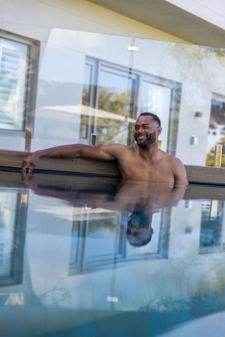 African american man relaxing in modern home pool with reflective water and glass terrace