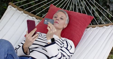 Mature woman relaxing in hammock using smartphone and credit card for mobile shopping