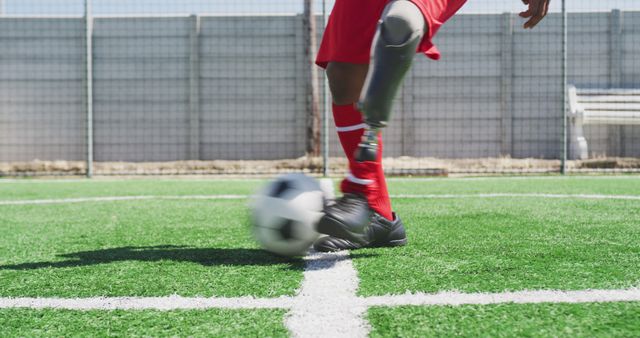 This photo shows a soccer player wearing a red uniform with a prosthetic leg while kicking a black and white soccer ball on a grass field. It can be used to promote inclusivity in sports, represent athletes with disabilities, or highlight the resilience and determination of individuals overcoming physical challenges.