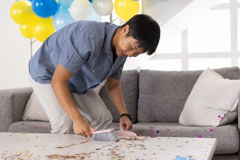 Asian man sweeping party confetti in sunny living room with balloons and gray sofa