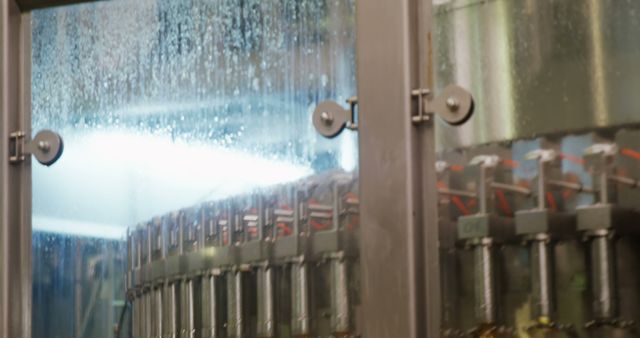 Industrial bottling machine seen through condensation on glass, filling beverages in glass bottles. Ideal for illustrating articles or advertising materials on manufacturing process, beverage production, industrial automation, or factory environments.