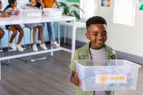 African american boy holding plastic recycling bin while classmates sorting recyclables