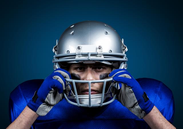 This image shows an American football player in a blue uniform adjusting his helmet, set against a blue background. The intense and focused expression highlights the competitive and determined nature of athletes in this sport. Ideal for use in sports-related articles, motivational posters, promotional materials for football games or training programs, and websites focusing on athletics or team sports.