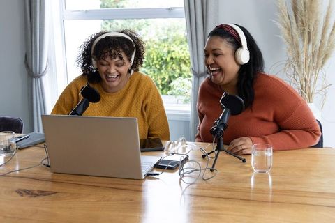 African american and indian women laughing, recording podcast together in home studio