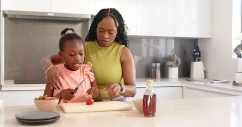 African american mother teaching daughter to cut strawberries on modern kitchen island