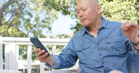 Asian senior man checking smartphone while sitting on porch chair in denim shirt outdoors