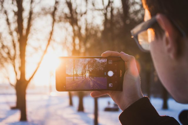 Person holding a smartphone capturing a scenic sunset in a snowy park. Bright sunbeams create a warm and enchanting atmosphere despite the cold season. Ideal for themes related to technology, winter, nature photography, and outdoor activities.