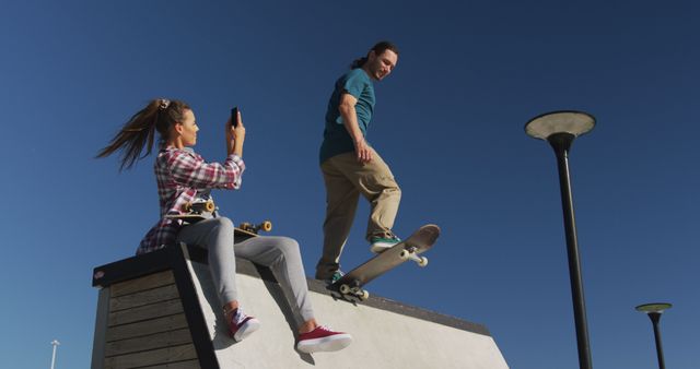 Two young adults engaged in skateboarding activities. Female skateboarder seated on ramp, recording male skateboarder performing tricks under clear blue sky. Ideal for use in sports lifestyle blogs, urban culture articles, and active lifestyle promotions.
