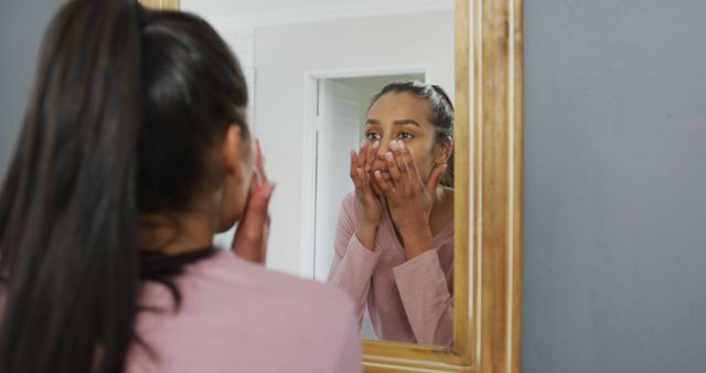 Young woman standing in front of mirror touching face and caring for skin. Ideal for advertising beauty products, skincare routines, self-care articles, or wellness blog posts.
