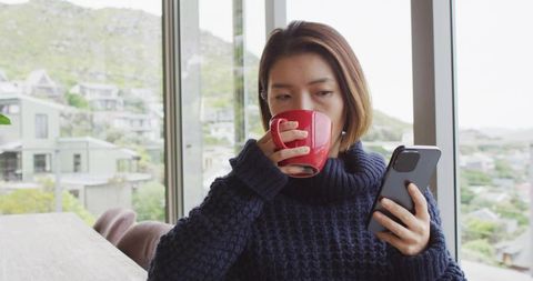 Sipping coffee while checking smartphone by window in cozy navy sweater, minimalist interior