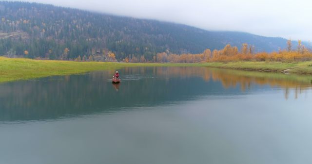 A person kayaks alone on a calm lake surrounded by vibrant autumn colors and a distant snowy mountain. The image captures the essence of tranquility and adventure, perfect for promoting outdoor activities, travel destinations, environmental awareness, and adventure tourism.