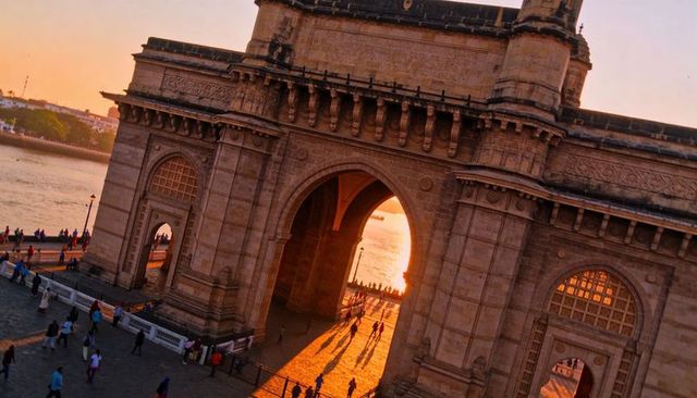 Gateway of india arch framing sunset over mumbai waterfront, people walking, golden shadows