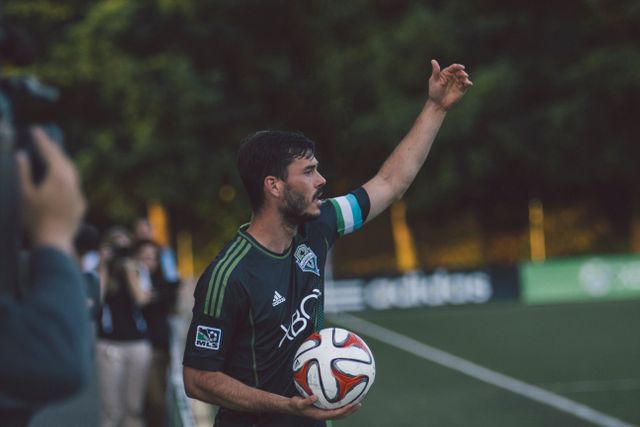 Soccer player in green jersey preparing to throw the ball back into play. Teammates and spectators seen in background. Perfect for illustrating concepts of sportsmanship, concentration, and athletic skill. Ideal for sports articles, training materials, and fitness promotions.