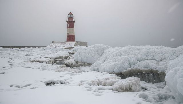 Red lighthouse guarding frozen breakwater amid snow-covered ice mounds on winter coastline