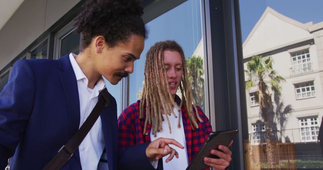 Two young urban professionals engage in a discussion while looking at a tablet. In an outdoor urban setting, they collaborate, highlighting the use of modern technology in business. Ideal for use in articles or advertisements related to business, technology, teamwork, or young professionals.