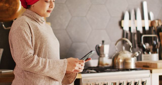 Middle-aged woman in a cozy kitchen setting with modern decor, holding and using a smartphone. She wears a red headscarf and a beige sweater. The background displays various cooking utensils and a teapot on the stove. Ideal for concepts such as domestic life, technology at home, staying connected, or modern interiors.