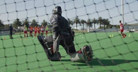 Kneeling field hockey goalkeeper blocking shot during outdoor training on green turf