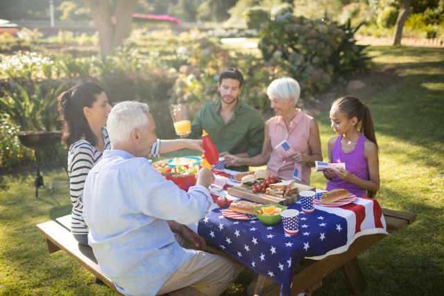 Multigenerational family enjoying a picnic in a park on a sunny day. They are sitting around a table covered with an American flag, sharing food and drinks. This image can be used for promoting family gatherings, outdoor activities, summer events, and patriotic celebrations.