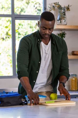 African american man packing healthy lunch at marble kitchen countertop with green apple
