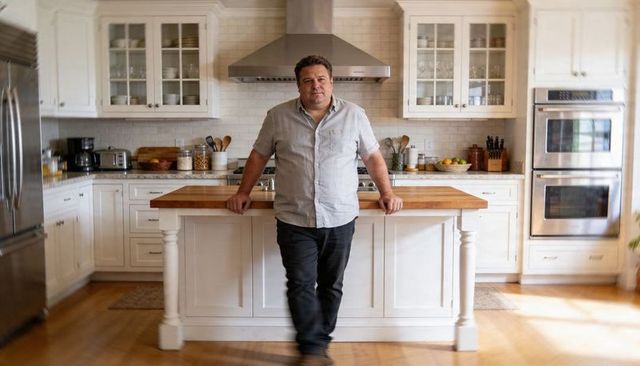 Man leaning on butcher-block island in sunlit farmhouse kitchen with stainless oven