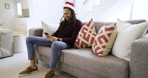 Dreadlocked man wearing santa hat relaxing on modern sofa checking smartphone