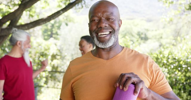 Middle-aged man holding a yoga mat and smiling outdoors, with friends in the background socializing. Surrounded by trees and a natural environment, the scene suggests a healthy and active lifestyle. Ideal for use in articles or advertisements related to fitness, wellness, outdoor activities, and social gatherings. Great for promoting positivity, community spirit, and the joys of staying active with friends.