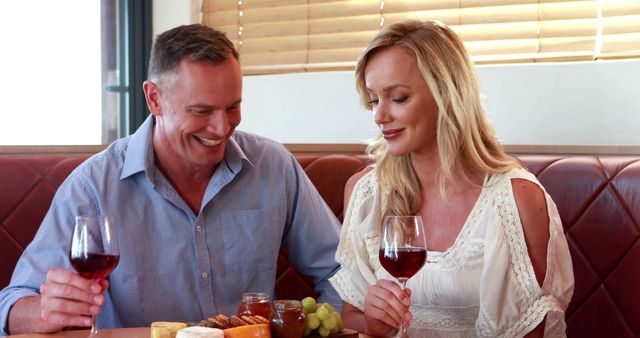 A middle-aged Caucasian couple enjoys a romantic moment with wine and snacks at a cozy dining setting, with copy space. Their cheerful interaction suggests a celebration or a special date, enhancing the intimate atmosphere.