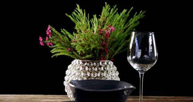 A vibrant green plant with pink flowers is displayed in a textured glass vase beside an empty wine glass and a black bowl on a wooden surface. The contrast between the colorful flora and the dark background creates a visually striking composition.