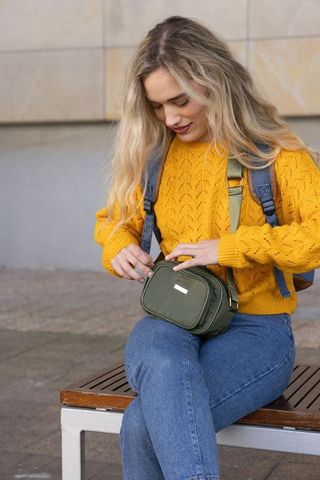 Young woman unzipping olive green crossbody bag on bench wearing mustard sweater casual commuter