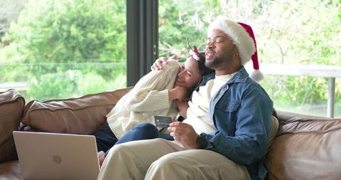 Couple wearing santa hat cuddling while online holiday shopping with credit card, laptop
