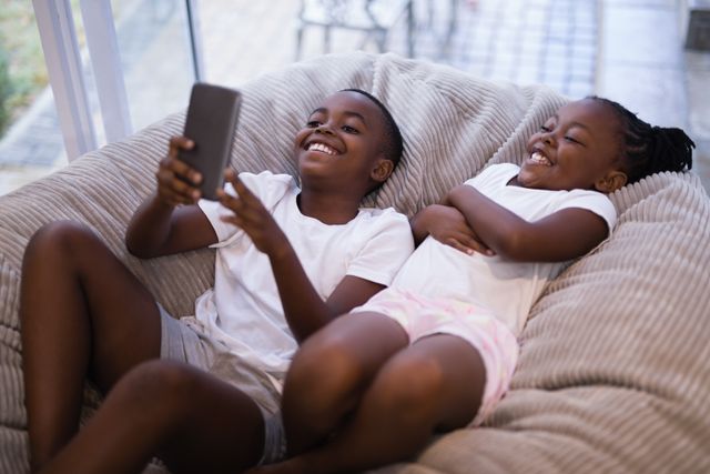 Happy siblings using mobile phone while lying on couch at home