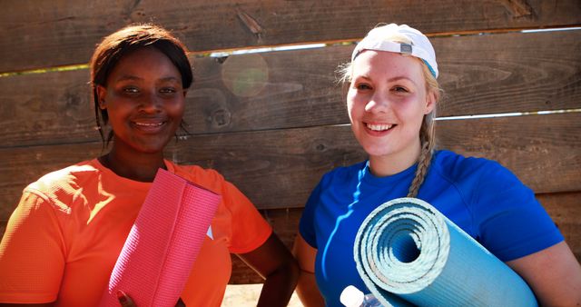 Two women, one in an orange shirt and the other in blue, holding yoga mats while smiling outdoors. They appear active and fit, reflecting a healthy and active lifestyle. This can be used in fitness, wellness, diversity, and healthy living contexts.