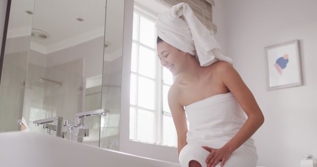 Image of smiling biracial woman with towel on hair sitting on bathtub in bathroom. Health and beauty, leisure time, domestic life and lifestyle concept.